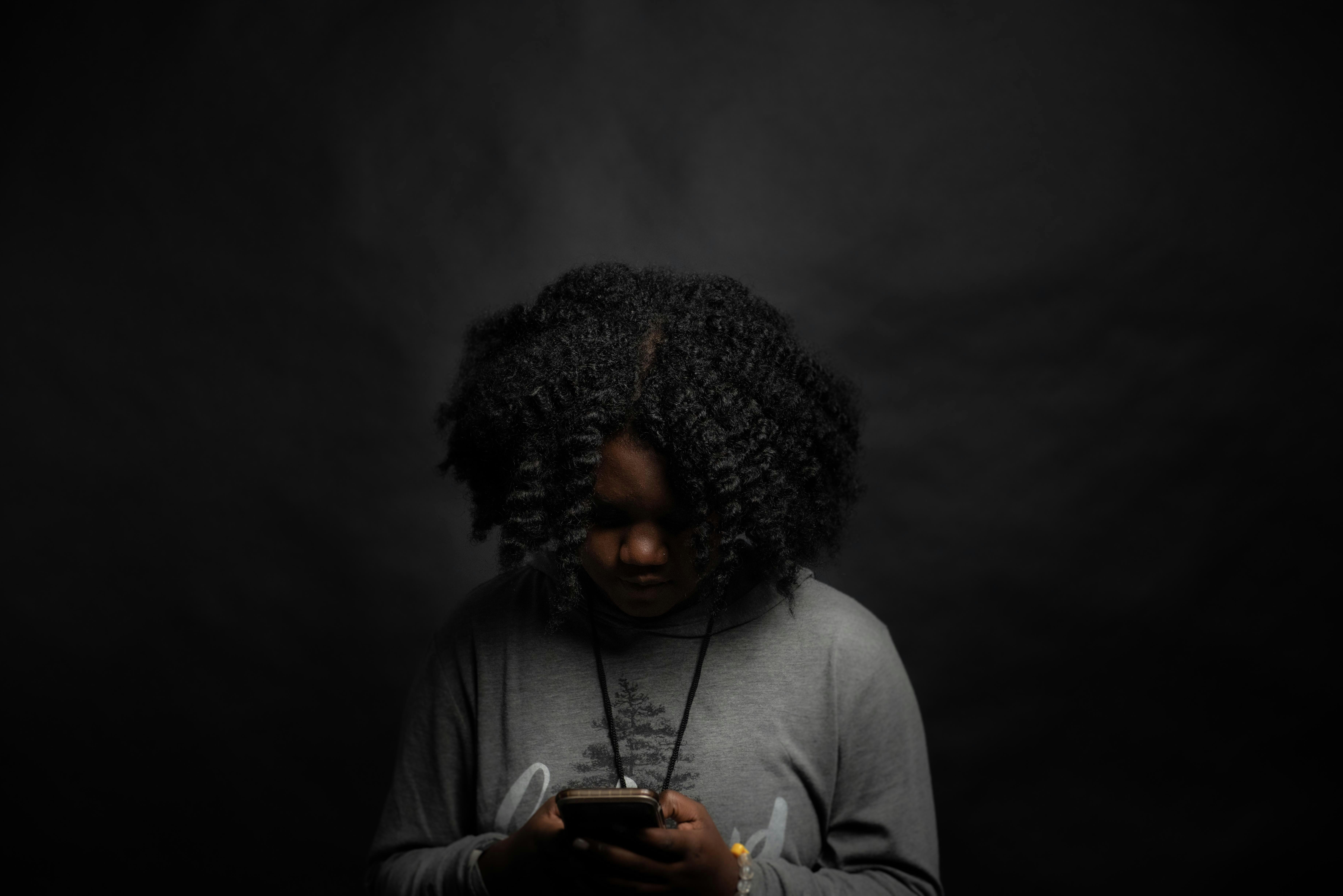 Young woman looking down at her phone in a dimly lit room, symbolizing the emotional weight and anxiety linked to social media